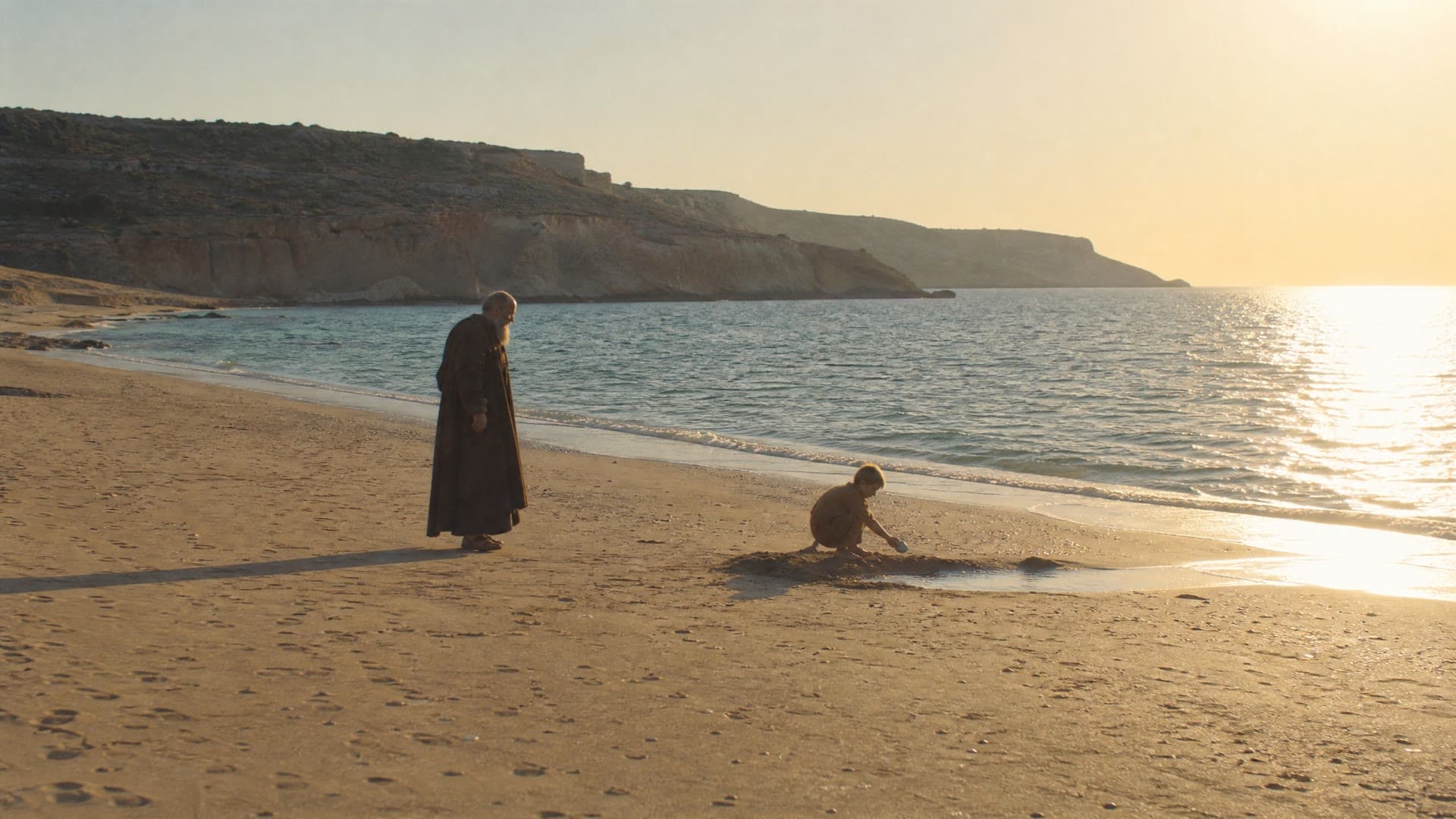 An old man in a dark robe watches a boy dig in the sand at the water’s edge, a cinematic reconstruction of a legend about Augustine